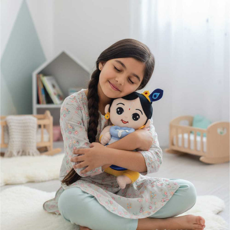 Young girl hugging a mantra chanting Krishna plush toy in a cozy room with a bookshelf and crib in the background.